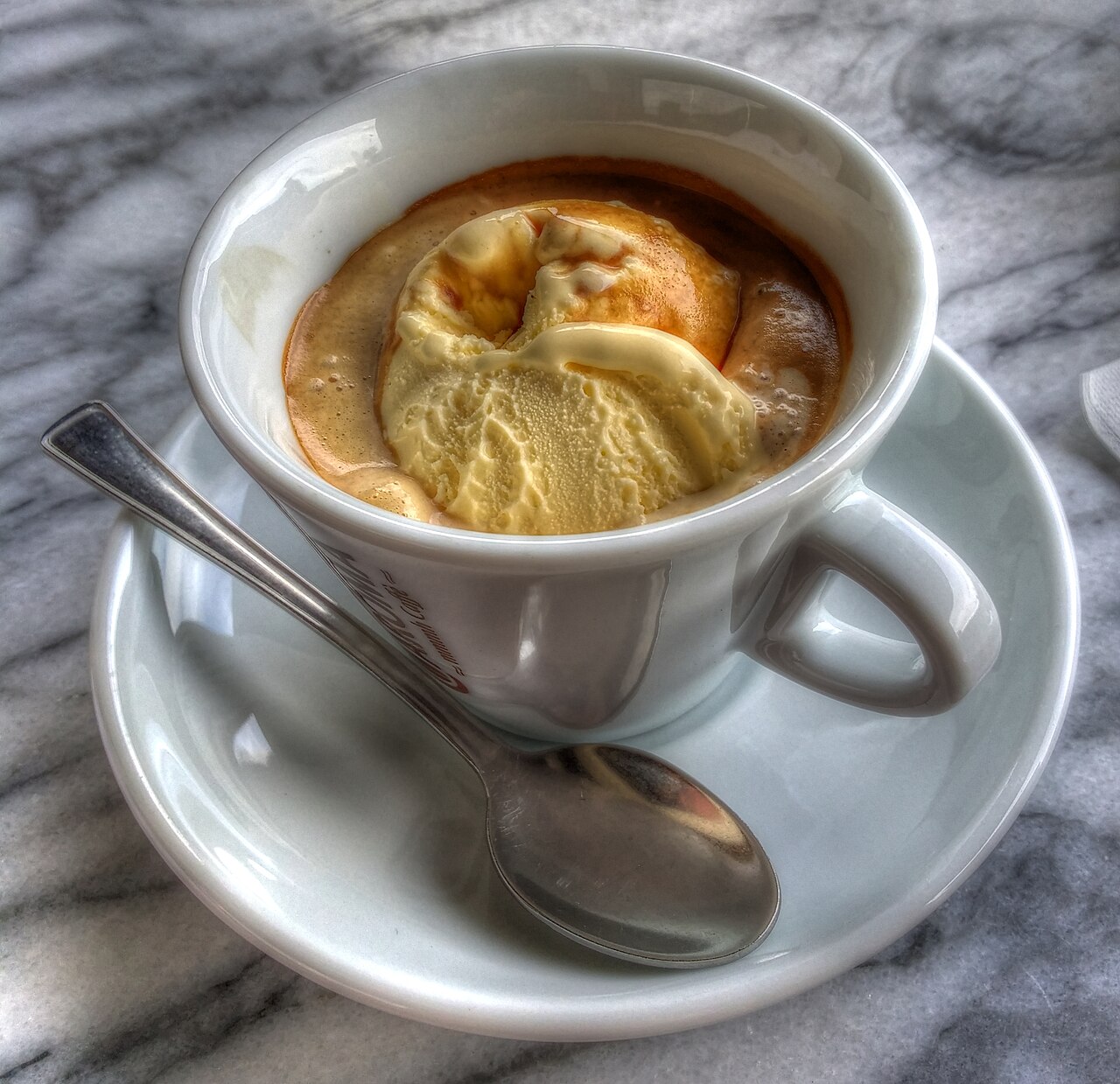 White cup containing an ice-cream scoop surrounded by espresso, with a spoon on its saucer.