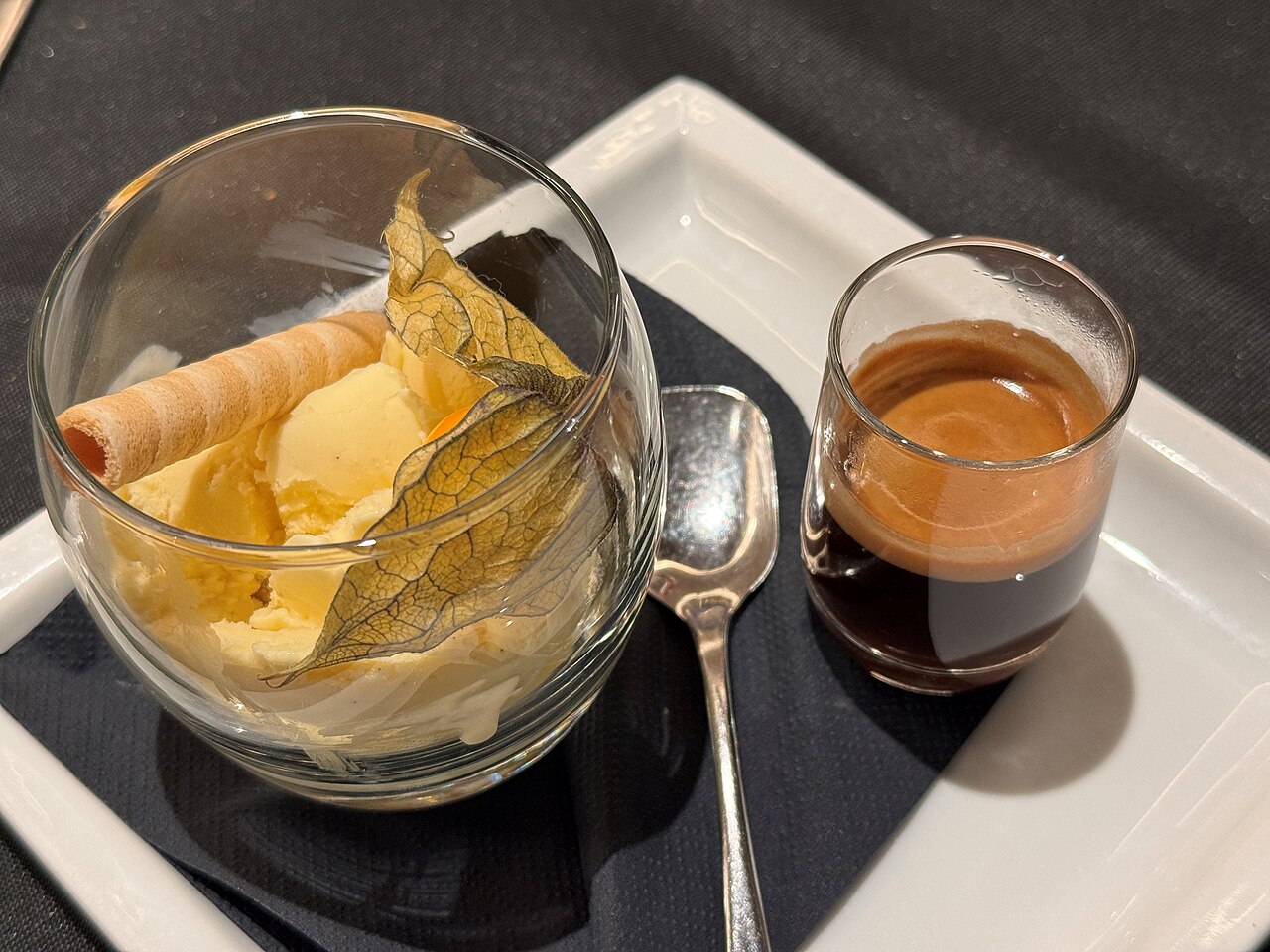 Ice cream in a clear bowl beside a separate espresso glass, with a spoon, wafer and fruit garnish.