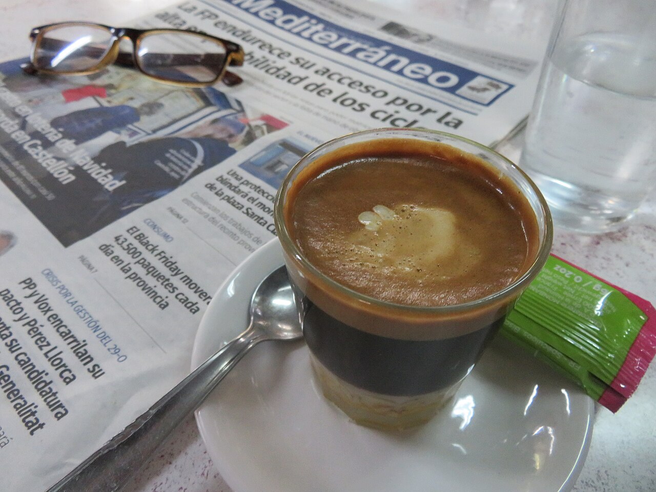 Small clear glass of café bombón with a pale bottom layer and dark coffee above, on a saucer.