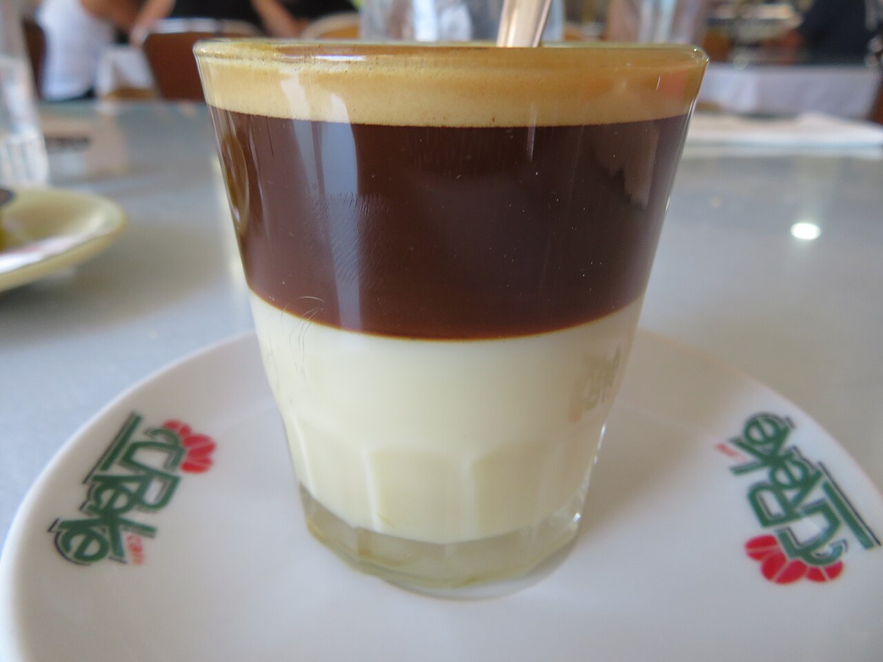 Clear glass with pale condensed milk below dark coffee and crema, with spoon and white saucer.
