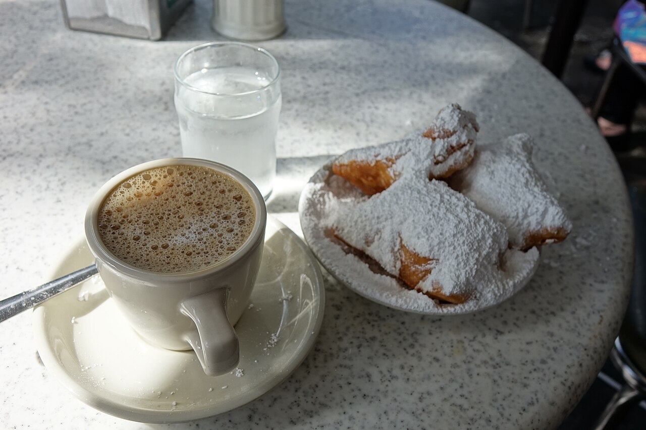 White cup of café au lait beside a plate of powdered-sugar beignets and a water glass.