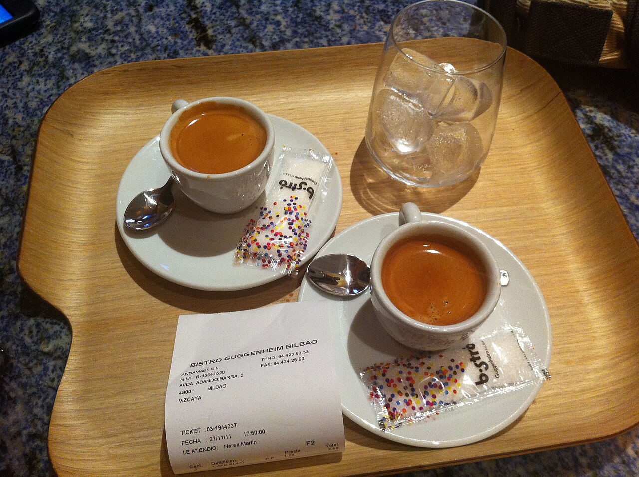 Tray with two cups of black coffee and a separate clear glass of ice, with spoons and sugar packets.