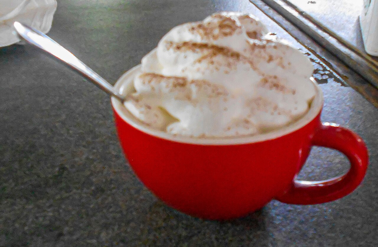 Red coffee cup with a generous whipped cream topping and a spoon on a café table.