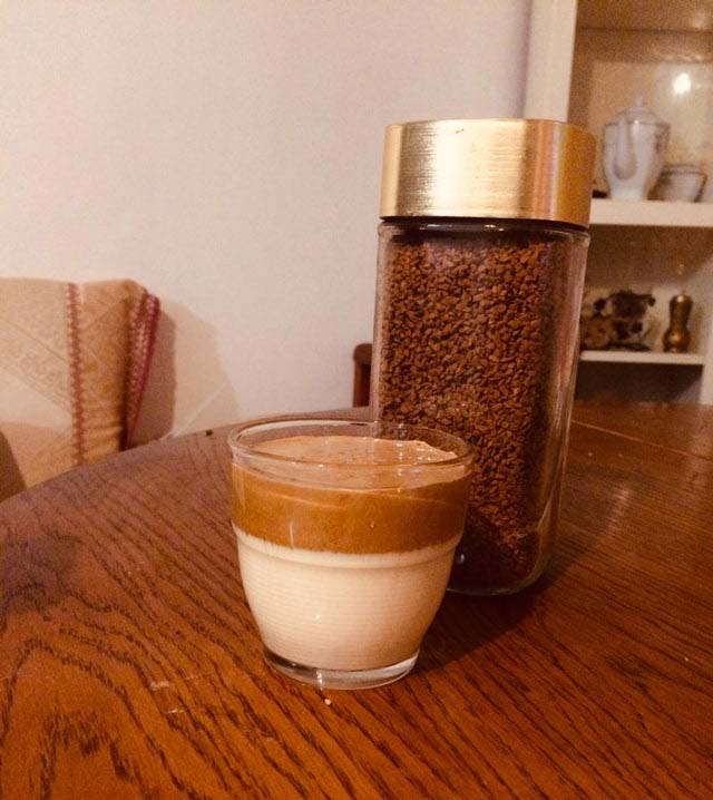 Small glass with pale milk below a brown whipped-coffee layer, beside an instant-coffee jar.