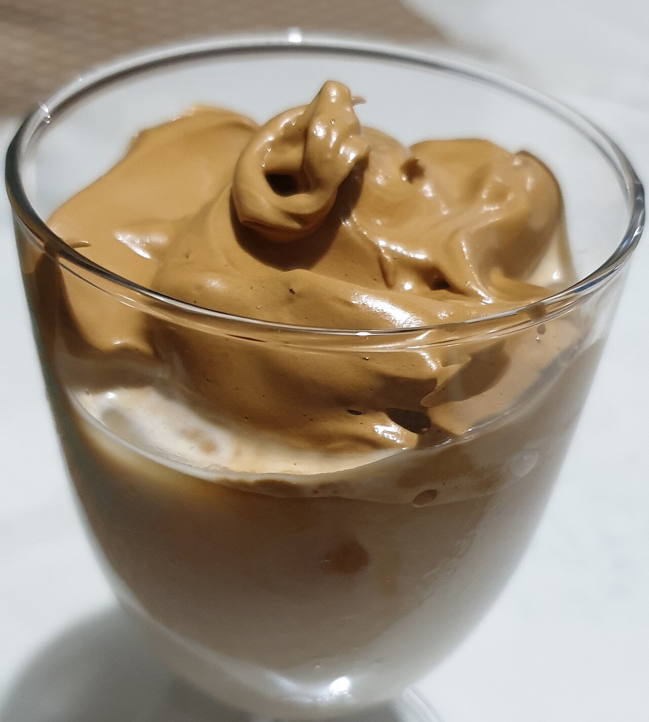 Clear glass of pale milk under a thick glossy brown whipped-coffee topping.
