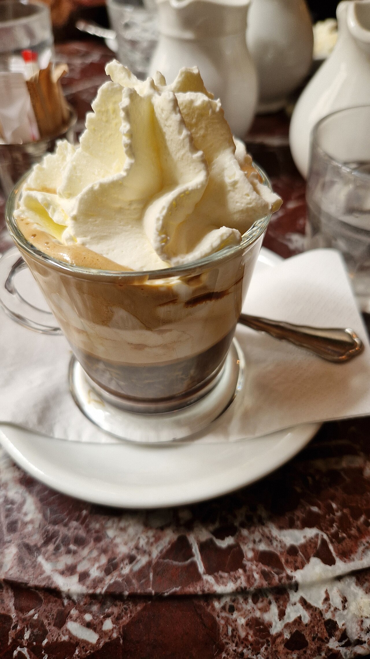 Handled glass of dark coffee with cream streaks and a large whipped-cream topping on a white saucer.