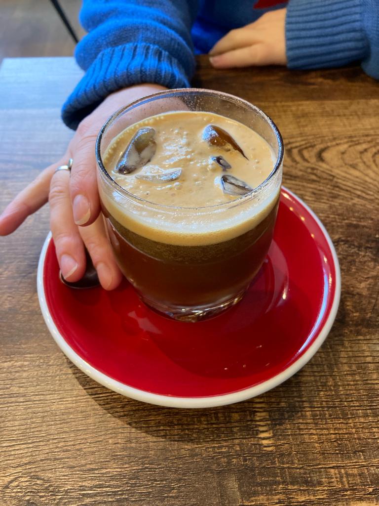 Clear tumbler of dark espresso tonic with ice and tan foam on a red saucer.