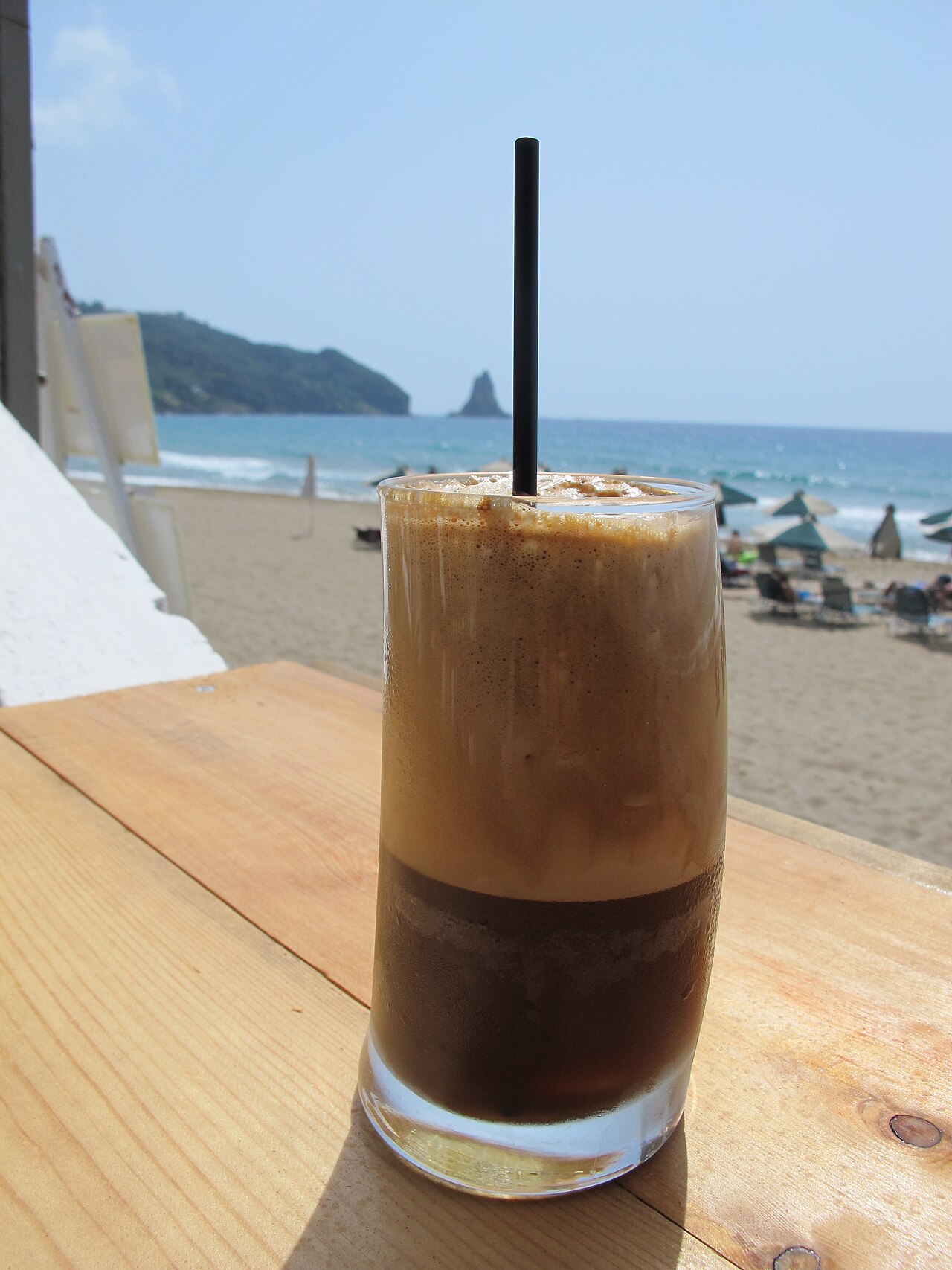 Tall clear glass of brown frappé coffee with a thick tan foam head and a straw.
