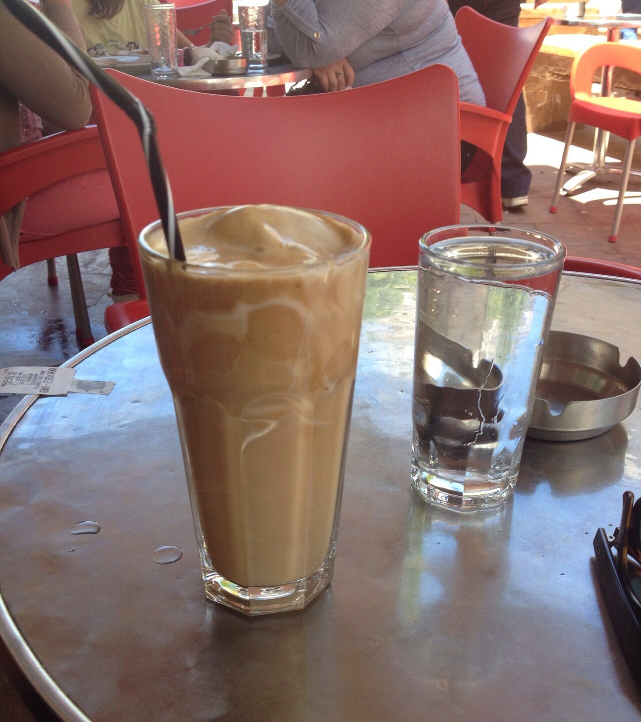 Tall clear glass of pale brown frappé coffee with a dark straw, beside a water glass.
