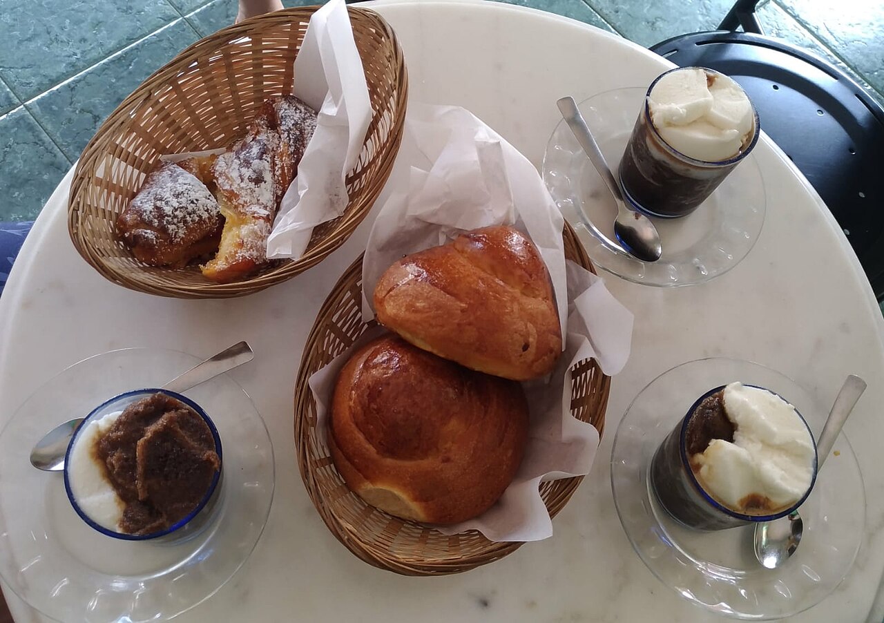 Small cup of dark and pale coffee-and-almond granita with a spoon, beside brioche and a cornetto.