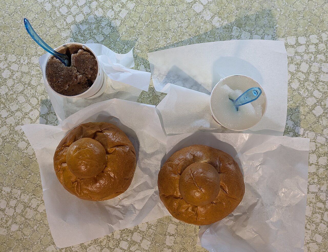 Two paper cups of granita, dark coffee at left and pale lemon at right, above two brioche rolls.