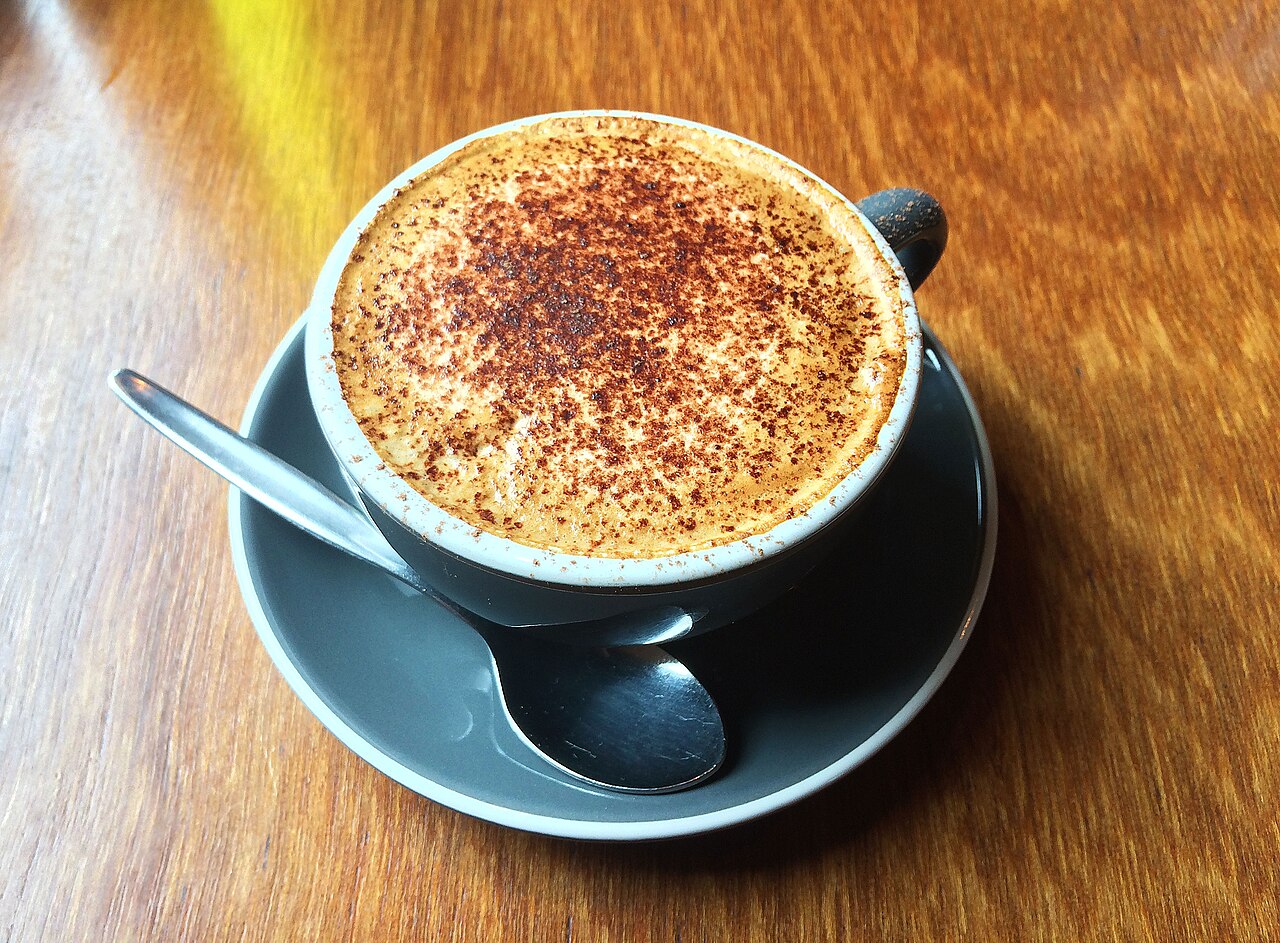Dark cup of foamy mocha with brown powder on its surface, on a saucer with a spoon.
