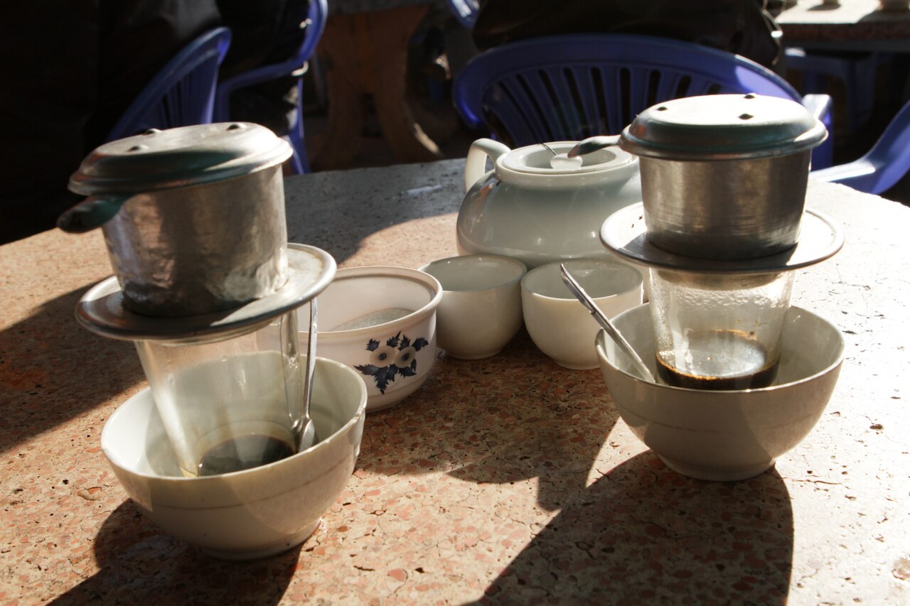 Metal phin over a clear glass with coffee and condensed milk, the glass resting in a bowl with tea beside it.