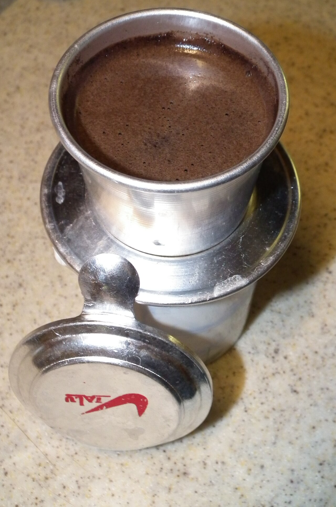 Metal phin filled with wet coffee above a receiving vessel, with lid and spoon beside.