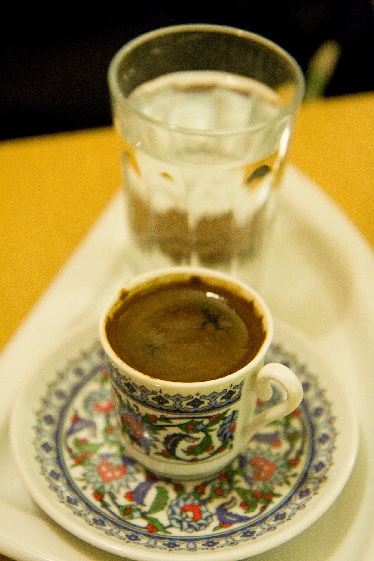 Small patterned cup and saucer holding dark Turkish coffee, with a water glass behind.